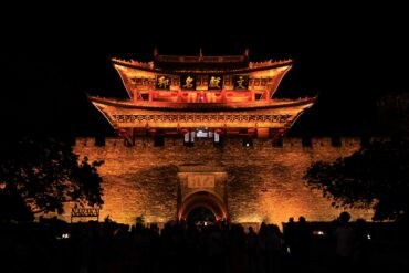 Dali Ancient City,Dali,Chinatour Night view of a lit ancient Chinese gate on a stone city wall with red lanterns and intricate carvings, silhouettes of people in the foreground