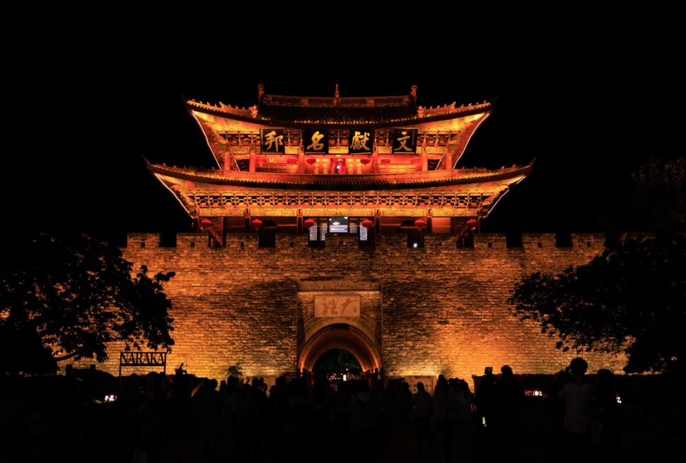 Night view of a lit ancient Chinese gate on a stone city wall with red lanterns and intricate carvings, silhouettes of people in the foreground