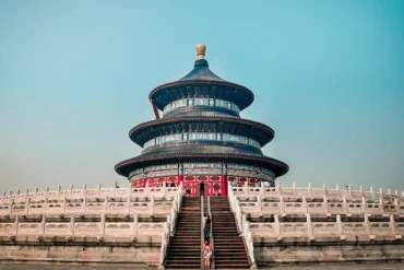 Circular, multi-tiered blue-tiled Chinese temple on a raised platform with white stone balustrades and central staircase, visitors entering.