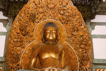 Golden Buddha statue seated in meditation, set against an intricately carved circular wooden halo in a temple alcove.