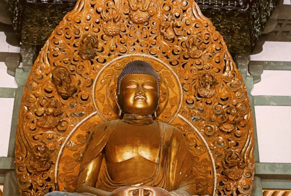 Golden Buddha statue seated in meditation, set against an intricately carved circular wooden halo in a temple alcove.