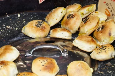 Round golden-brown buns surrounding a shiny metal lid on a dark baking surface, some topped with sesame seeds.