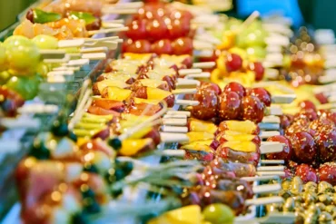Colorful fruit skewers arranged in a market display, glossy glazed pieces in red, yellow, and green on wooden sticks