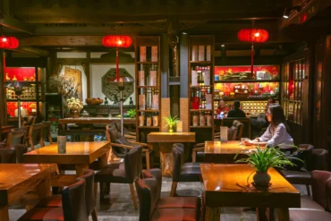 Dimly lit Asian restaurant interior with red lanterns, wooden tables and chairs, and shelves of decor in the background. A woman sits at a table on the right side of the image.