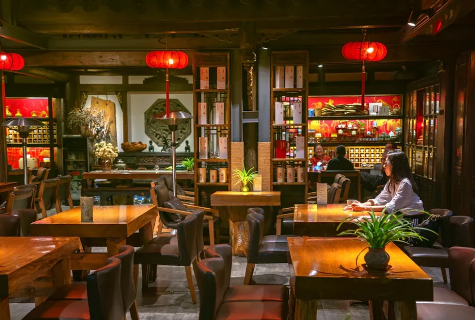 Dimly lit Asian restaurant interior with red lanterns, wooden tables and chairs, and shelves of decor in the background. A woman sits at a table on the right side of the image.