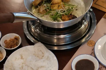Hot pot simmering on a portable burner with greens and meat, surrounded by sauce bowls, fried onions, and peanuts, plus a plate of white rice noodles.