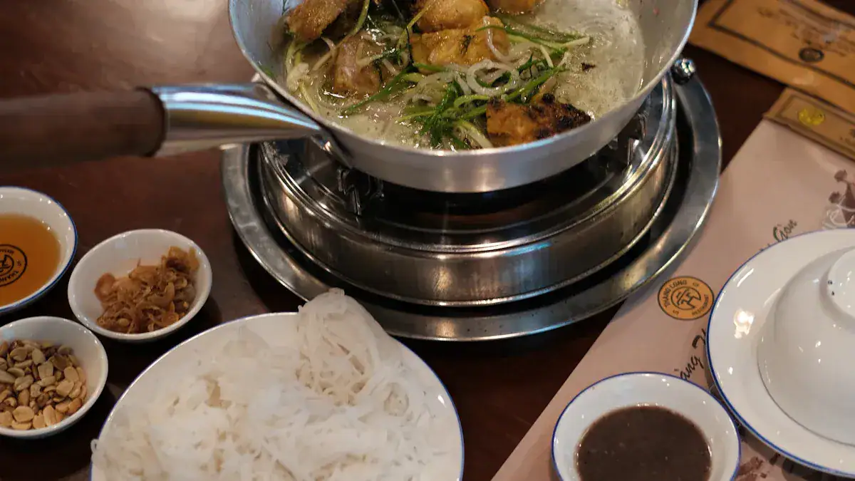 Hot pot simmering on a portable burner with greens and meat, surrounded by sauce bowls, fried onions, and peanuts, plus a plate of white rice noodles.