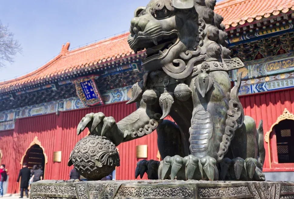 Stone dragon statue guarding a carved orb in front of a red traditional Chinese temple with ornate multicolored eaves and visitors in the background.