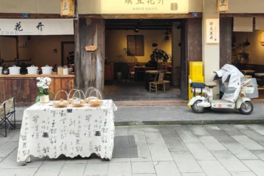 Open storefronts on a quaint street with a table of pastries covered by a white cloth in front and a delivery scooter parked to the right.