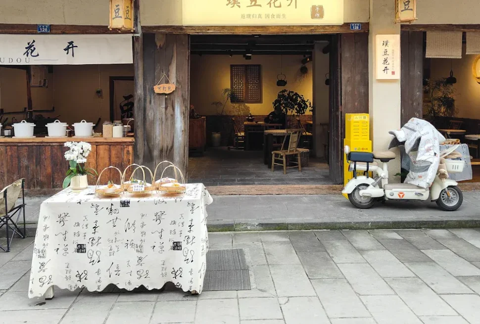 Open storefronts on a quaint street with a table of pastries covered by a white cloth in front and a delivery scooter parked to the right.