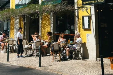 Outdoor sidewalk café with patrons seated at wicker tables in front of a bright yellow building labeled 'Asian Street Food'.