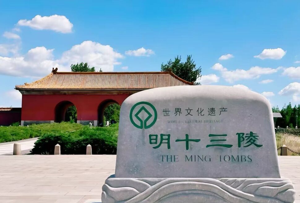 Ming Tombs,Beijing,Chinatour Stone monument with green emblem and Chinese text reading 'World Cultural Heritage' and 'The Ming Tombs' in front of a red gate with tiled roof under a blue sky.