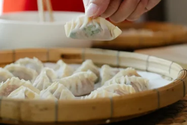 Bamboo steamer filled with steamed dumplings as a hand lifts one up with chopsticks.