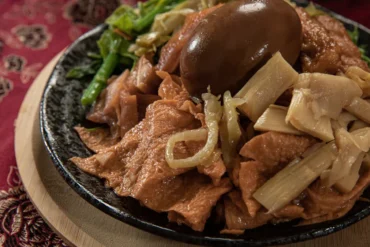 Sliced beef in a dark sauce with onions and bamboo shoots in a black skillet, garnished with greens on a red tablecloth.