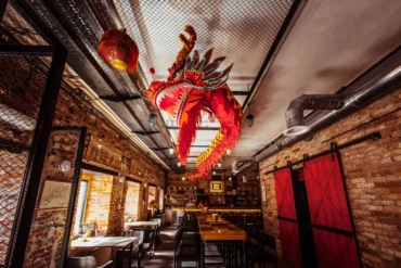 Long communal dining area with brick walls and a vivid red dragon sculpture hanging from the ceiling above the tables.