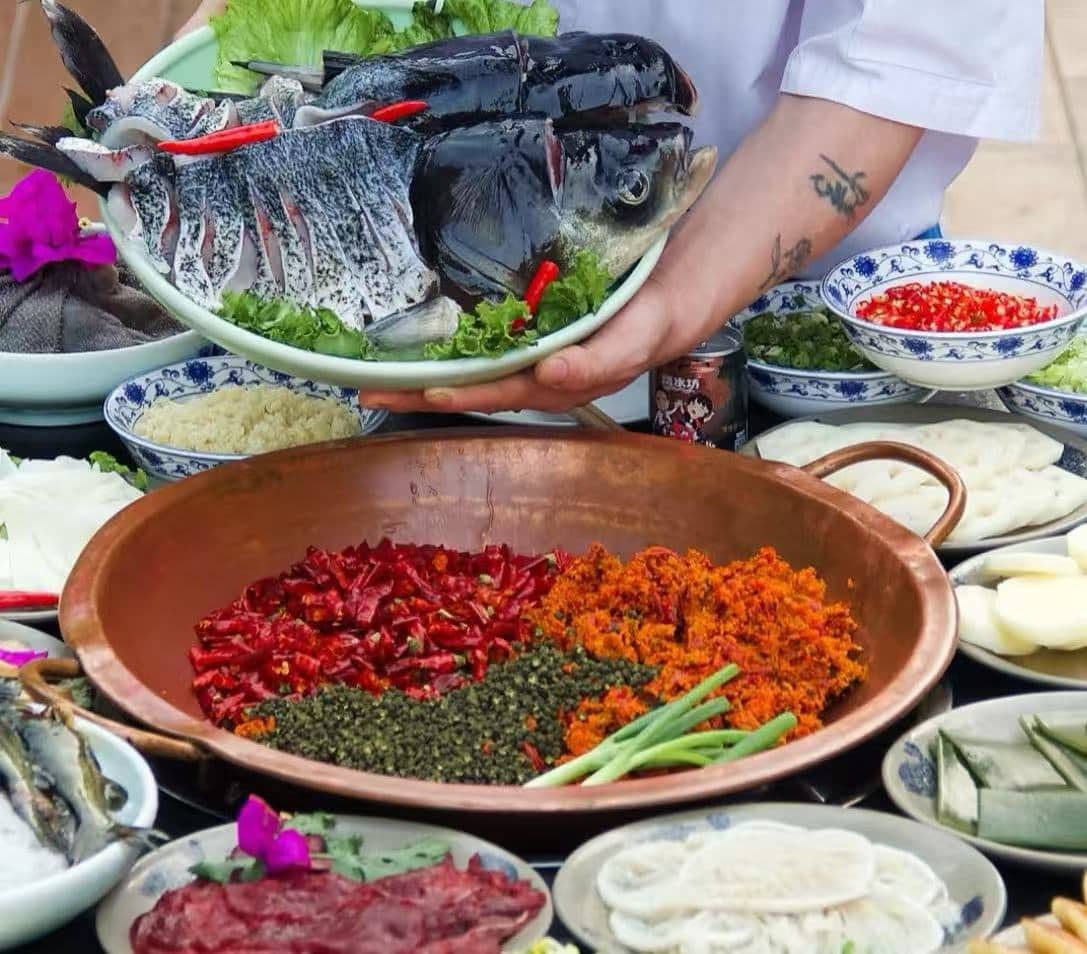 A person holds a plate of whole fish heads above a table of ingredients; in a copper bowl in the foreground are chopped red chilies, orange spices, green herbs, and scallions surrounded by bowls of condiments.
