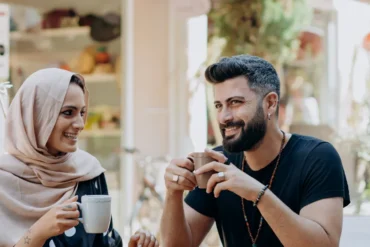 Two friends enjoy coffee at an outdoor cafe, smiling at each other as they hold mugs.
