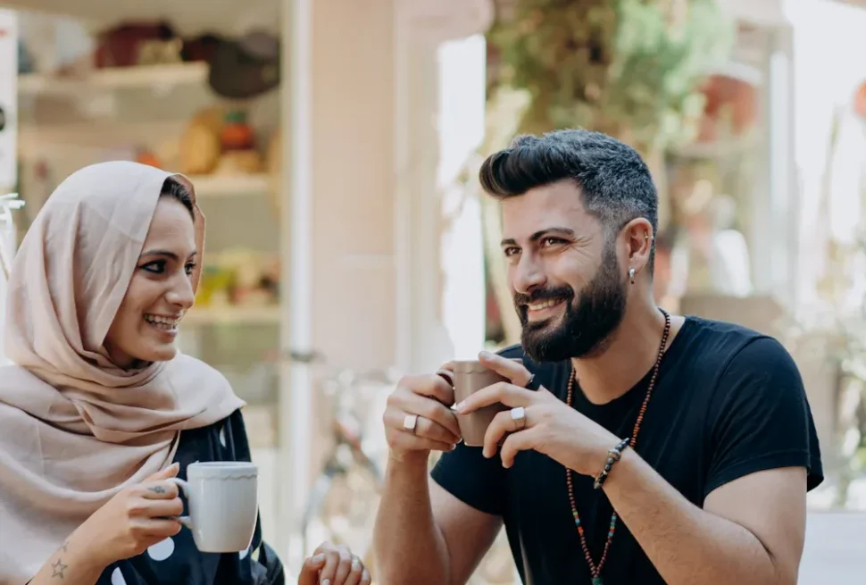 Two friends enjoy coffee at an outdoor cafe, smiling at each other as they hold mugs.