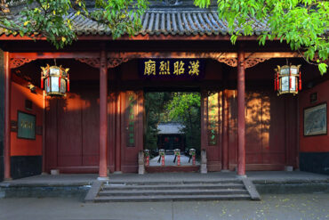 Red wooden Chinese temple gate with hanging lanterns and a courtyard beyond, under green trees