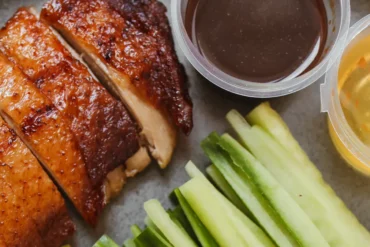 Sliced roasted chicken with crispy skin beside cucumber sticks and two dipping sauces in clear cups