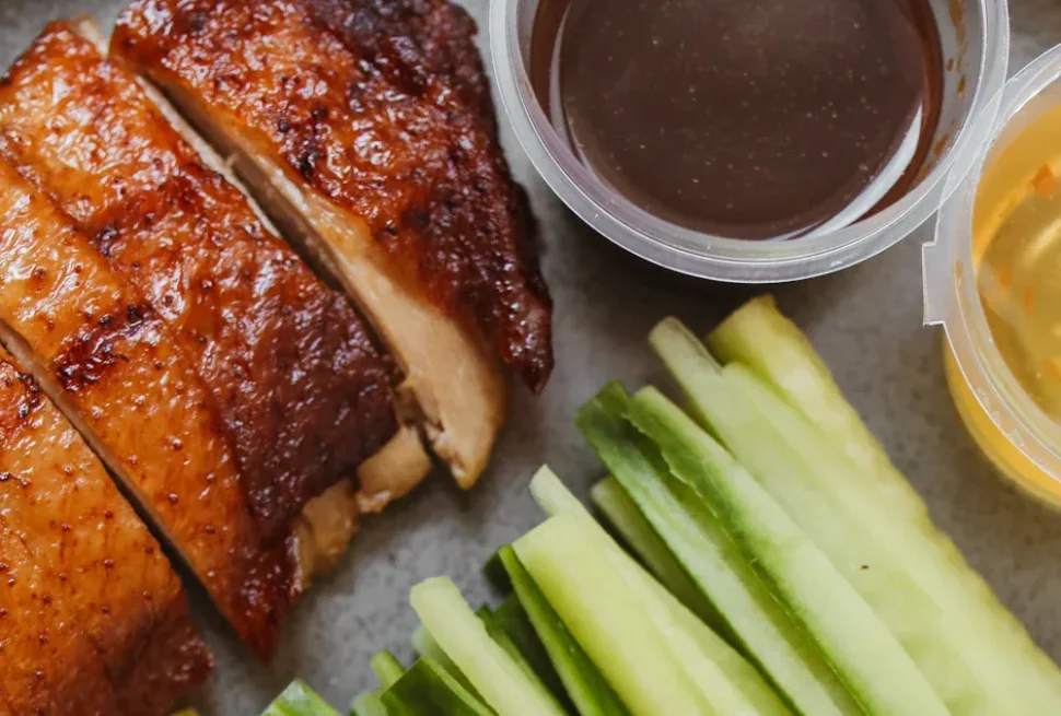 Sliced roasted chicken with crispy skin beside cucumber sticks and two dipping sauces in clear cups