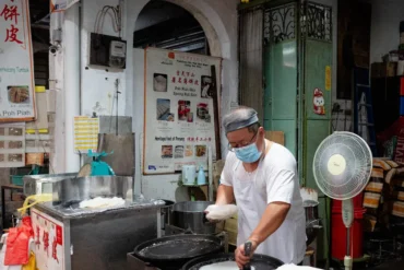 Man wearing a mask and headband cooks at a street food stall, using a large pan with equipment and signs in the background.