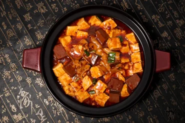 Mapo tofu in a black pot with red chili sauce, cubed tofu and green onions over a dark patterned surface with Chinese writing