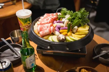 Hot pot with sliced pork, vegetables, and greens on a tabletop burner, with drinks and bowls nearby on the table.