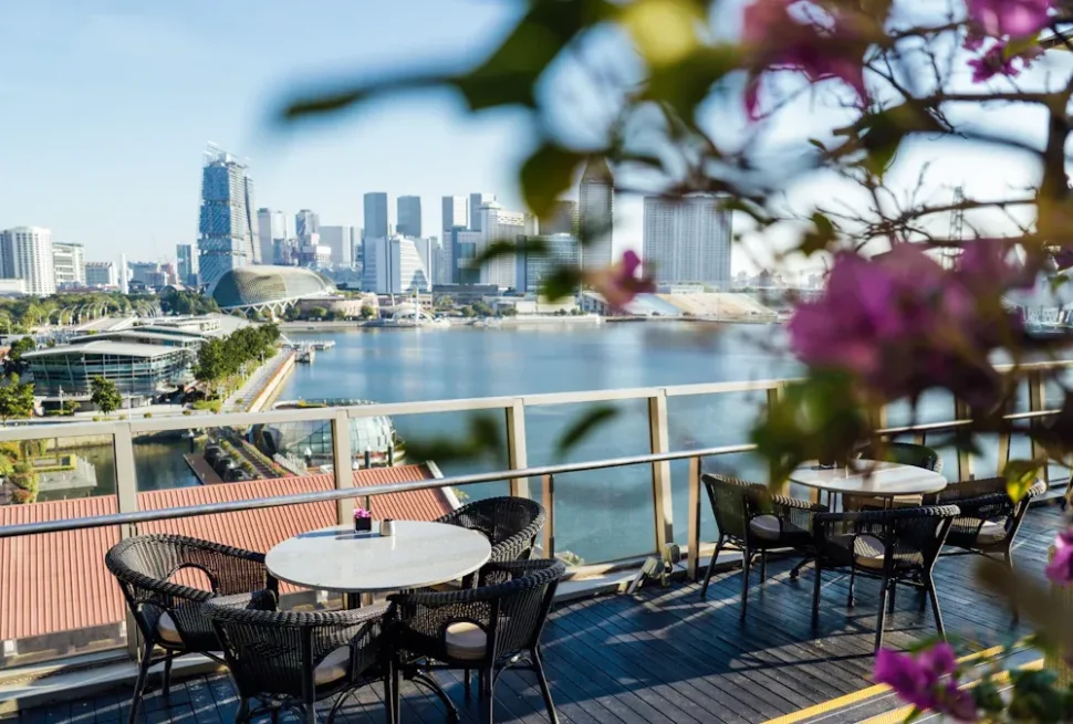 Rooftop terrace with round marble tables and black wicker chairs overlooking a marina and city skyline.