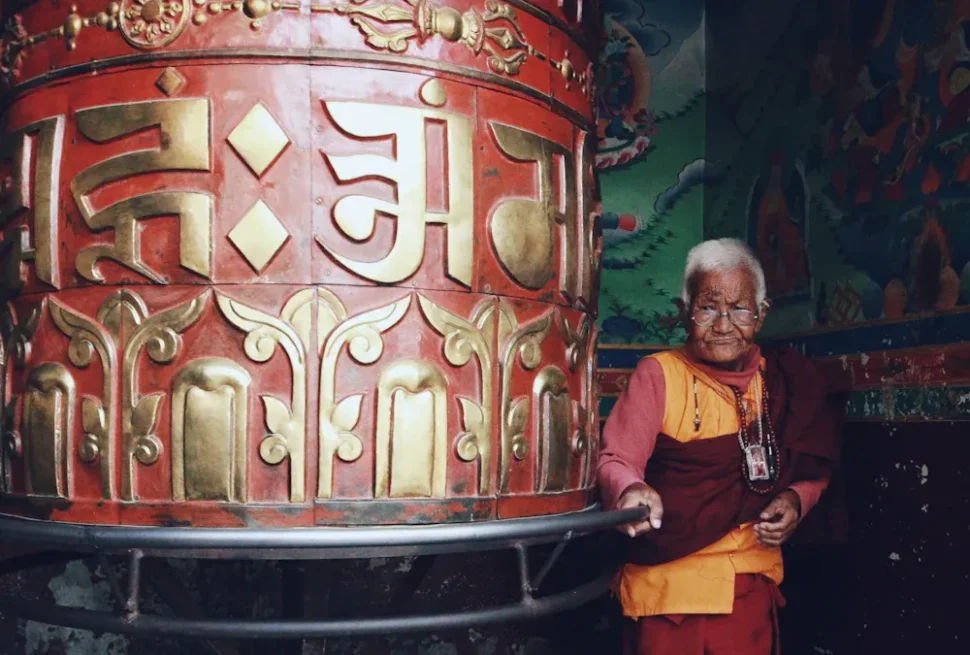 Elderly Buddhist monk in orange robes leaning on a large red and gold prayer wheel inside a colorful temple.