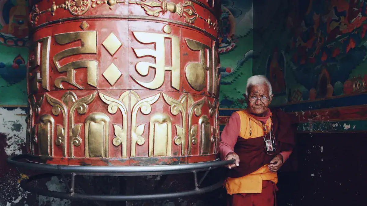 Elderly Buddhist monk in orange robes leaning on a large red and gold prayer wheel inside a colorful temple.