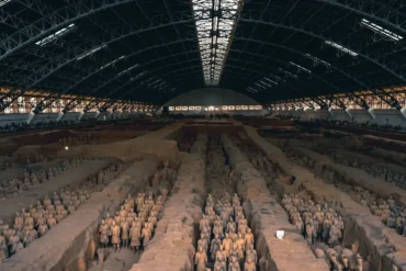 Expansive indoor excavation site showing rows of Terracotta Army soldiers in long trenches under a large arched roof.