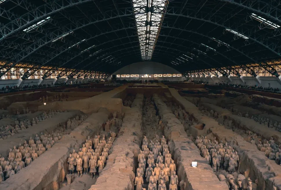 Expansive indoor excavation site showing rows of Terracotta Army soldiers in long trenches under a large arched roof.