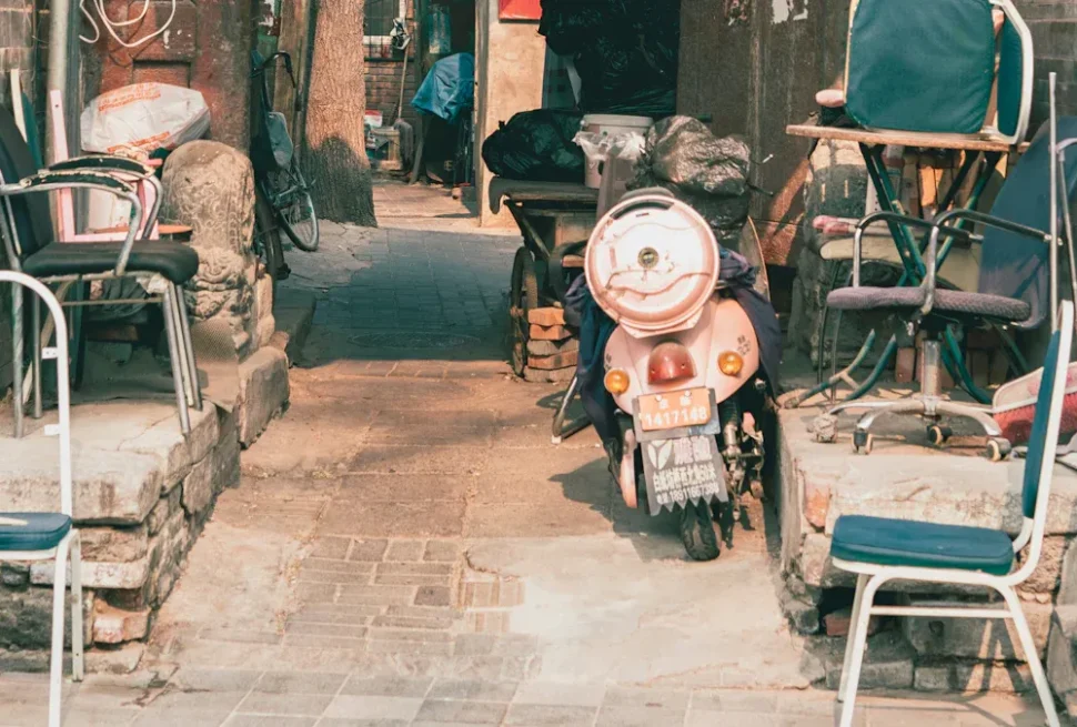Scooter parked in a narrow alley, flanked by chairs and scattered items, urban clutter all around.