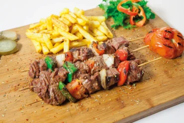 Beef kebab skewers with onions and peppers, served with fries and a grilled tomato on a wooden board