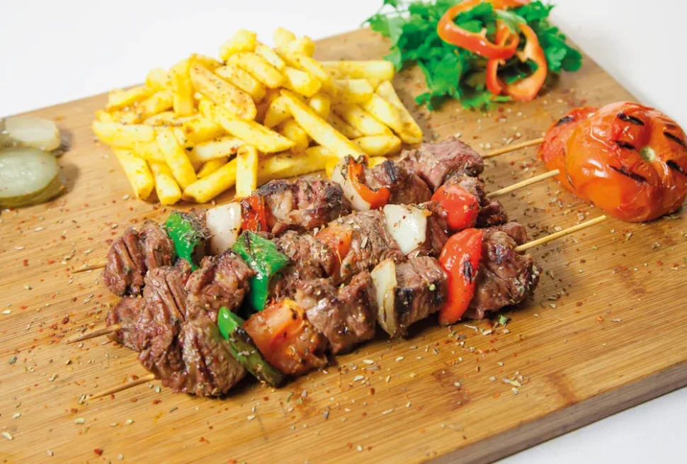 Beef kebab skewers with onions and peppers, served with fries and a grilled tomato on a wooden board