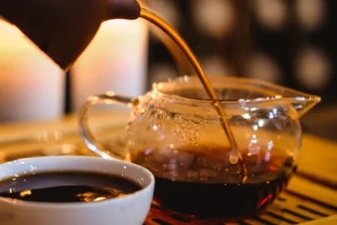 Pouring hot coffee from a kettle into a glass coffee pot with a white cup of black coffee nearby