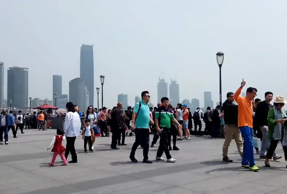 Crowded waterfront promenade with pedestrians and a distant city skyline of tall buildings on a clear day.