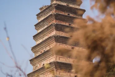 Brick, multi-tiered pagoda rising against a blue sky, with blurred foreground branches partially obscuring it.