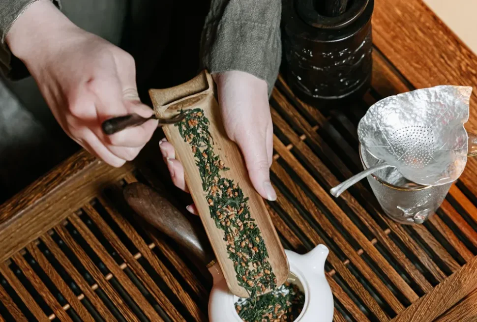 Hands crush dried herbal leaves on a wooden block using a small tool beside a teapot and metal strainer on a wooden tray.