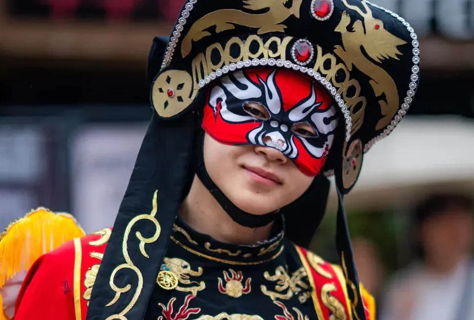 Person wearing an ornate black, gold, and jewel-encrusted headdress with a red and black decorative mask, likely a festival performer.