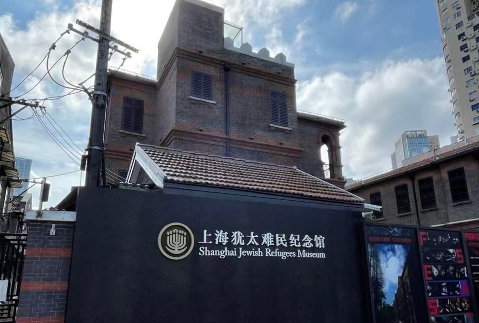 Exterior view of the Shanghai Jewish Refugees Museum with a dark wall sign displaying the museum name in Chinese and English behind a brick building.