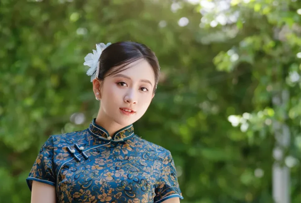 Shanghai Qipao Photo,Chinatour Young woman wearing a blue floral cheongsam outdoors, with a white flower in her hair, gazing softly at the camera.