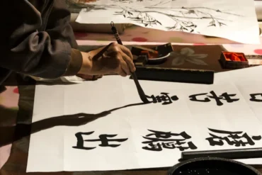 Close-up of a person’s hand holding a calligraphy brush, writing bold black characters on white paper with ink and an ink stone nearby.