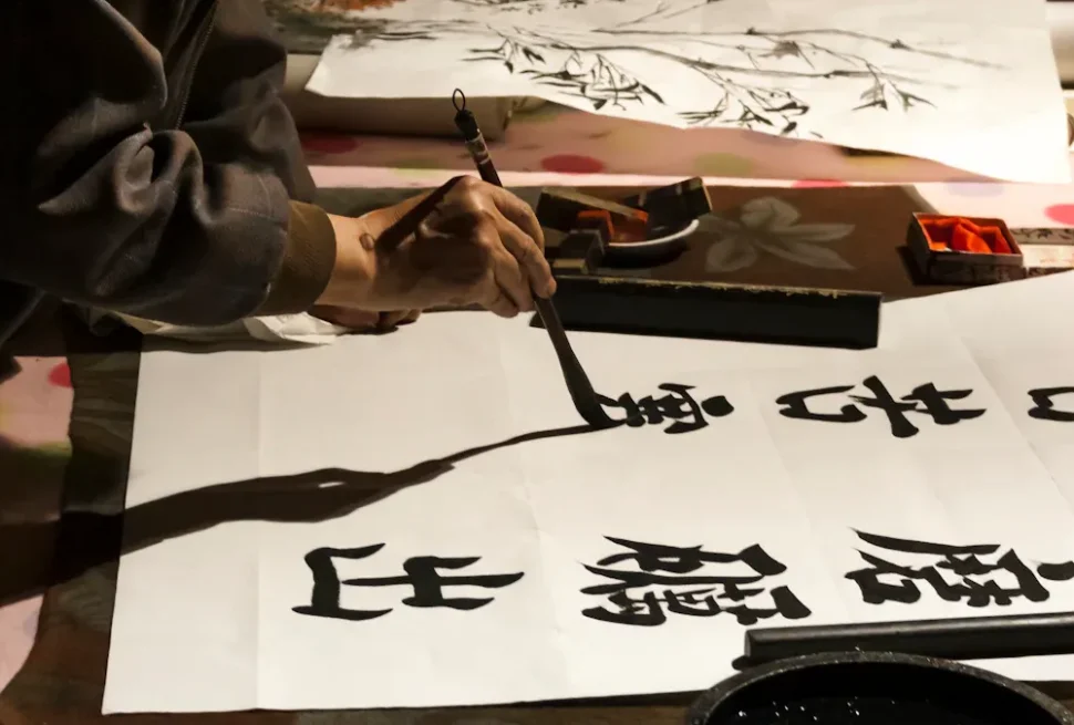 Close-up of a person’s hand holding a calligraphy brush, writing bold black characters on white paper with ink and an ink stone nearby.
