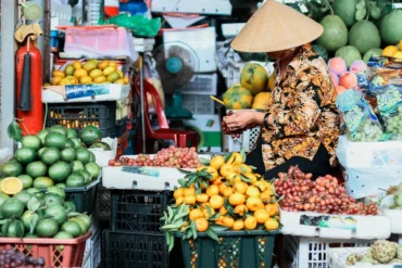 Market stall scene with a woman in a conical hat sorting oranges among crates of fruit at a busy outdoor market.