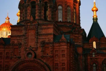 Red-brick church with ornate arches and a dark roof, gold-domed building visible behind it in the background.