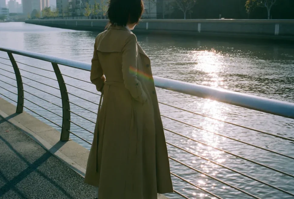 Person in a beige trench coat leans on a railing by the river, watching the sun glow on the water surface.
