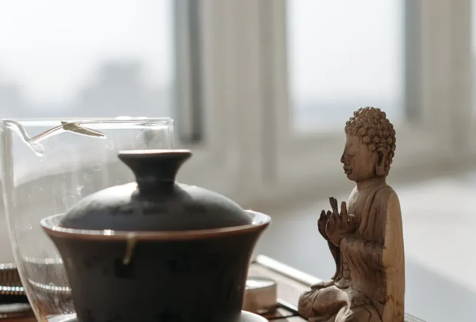 Tea Houses,Chinatour Wooden Buddha statue in meditation beside a dark ceramic teapot and a glass pitcher on a sunlit table by a window