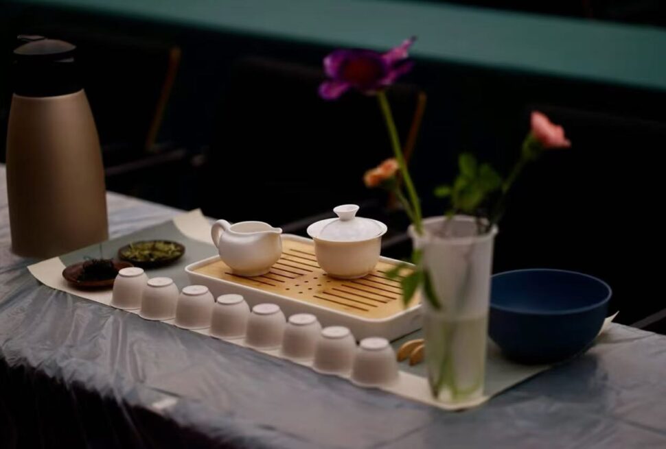 Shanghai Tea Ceremony,Shanghai,Chinatour White ceramic tea set on a tray with a teapot and a row of upside-down cups, on a table with a vase of pink flowers nearby.
